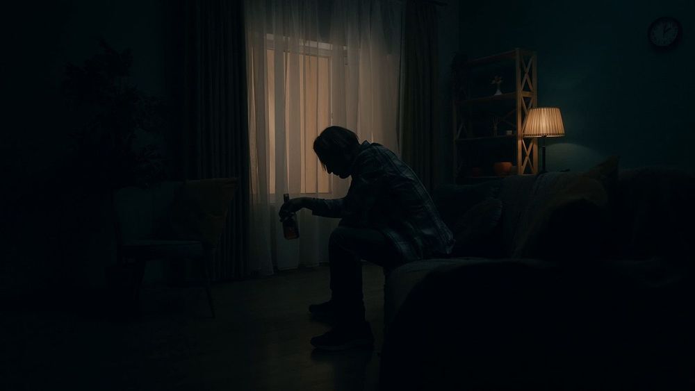 A devastated young man sits in a room in the dark with a bottle of alcohol. Alcoholism. Hopelessness. Alcohol addiction treatment. The concept of mental health.