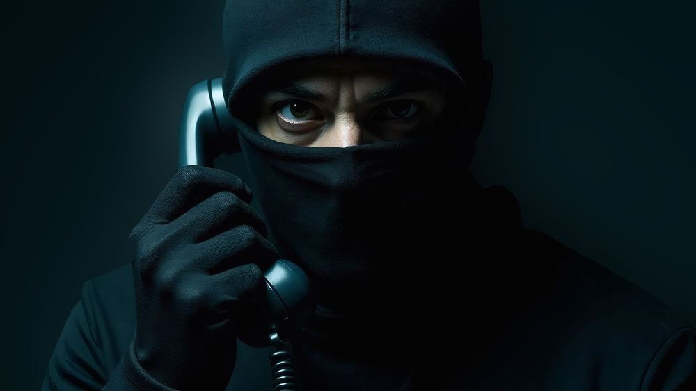 Male scammer wearing balaclava and mask, wearing black gloves, calling on phone in dim light. Anonymity, crime and fraud concept.