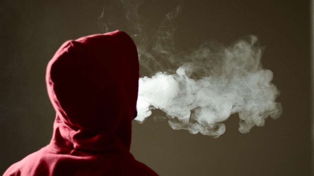 Young male in red hoodie vaping smoking, exhales thick vapor, is